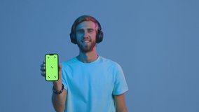 Music apps for phone. Modern young guy in headphones shows smartphone with template green screen, walks, enjoying sound