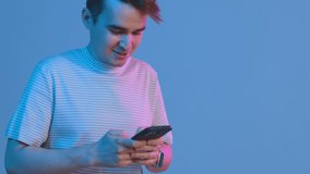Close up video of a young man in casual clothes walking in studio on blue background and texting a message. Guy smiling.