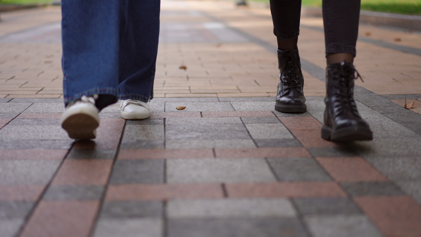 Front view steps of two unrecognizable women strolling along pavement alley in city park. Unrecognizable Caucasian friends walking outdoors. Lifestyle concept