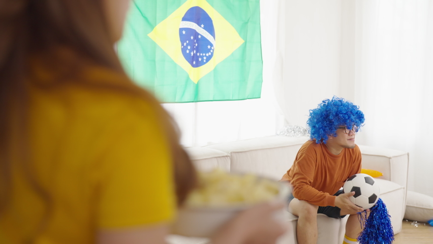 4K Asian man and woman sport fans shouting and celebrating football team victory the match during watching world soccer football competition on television with eating food and drinking beer at home.