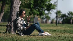 Modern Arabic guy talking online video call headphones laptop at summer tropical park with palm tree. Happy freelancer business student chatting videoconference e learning telecommuting teleworking