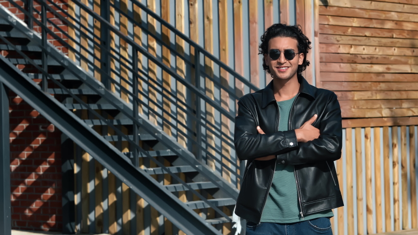 Trendy modern attractive man smiling downtown outdoor posing wooden building exterior with crossed hands. Handsome masculine young Arabic guy in black leather jacket relaxing urban city street