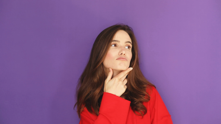 Pensive puzzled brunette young girl thinking isolated on purple background in studio. Girl put hand prop up on chin. Thoughtful woman, grimacing thinking girl, she look coquette and dreaming. Royalty-Free Stock Footage #1096645593