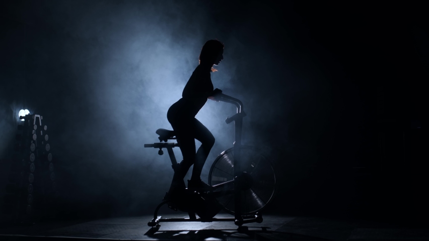 Athletic girl using exercise equipment in a modern dark gym. A young woman rides a bicycle in the smoke. Aggressively Muscular Pedaling on a Stationary Be in a Dark Gym. Healthy lifestyle Royalty-Free Stock Footage #1096647565