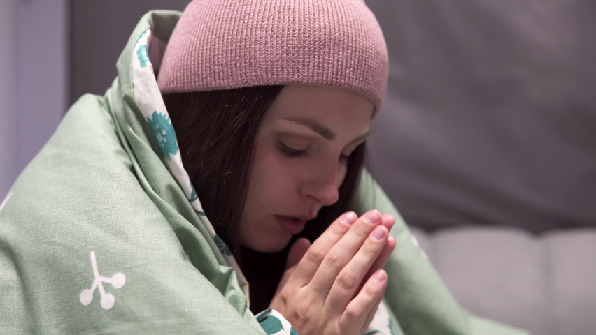 A young woman in a hat wrapped in a blanket is shivering from the cold on a bed in an unheated apartment. Shutdowns of electricity and central heating due to the crisis in the energy sector.