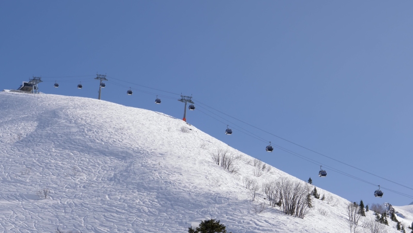 Ski resort on sunny winter day. Ski lift funicular cab lifts skiers and snowboarders up mountain against clear blue sky. Breathtaking view of snowy mountains resort and valleys of glaciers.