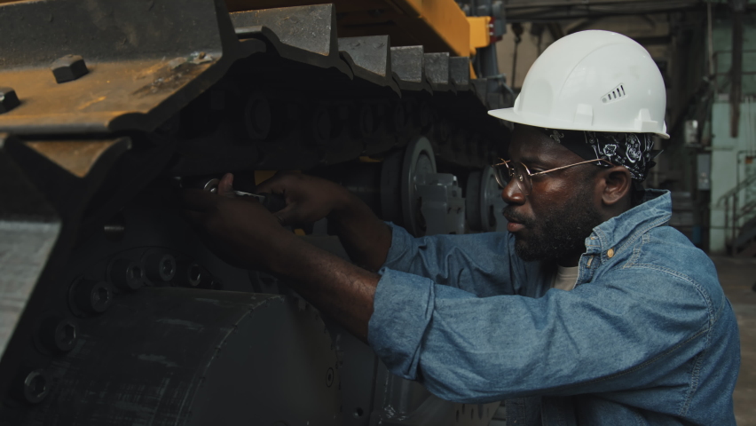 Hard working African American mechanic in denim shirt and hard hat using bolt wrench while fixing wheel loader at tractor plant | Shutterstock HD Video #1096652647