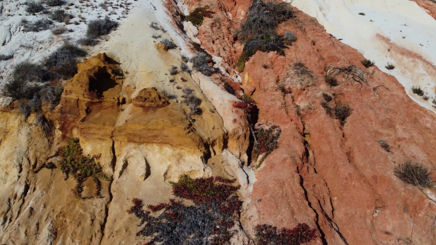 Drone view of an orange relief rock overgrown with various bushes. Sand avalanches descend along water-washed channels.
