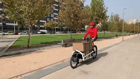 young messenger with red clothes and helmet riding a cargo bike to make a shipment on a bike lane in a city, horizontal video	
