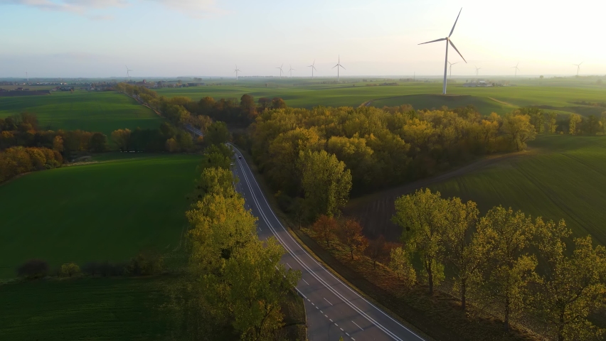 Aerial view above road near forest in full with cars in colorful countryside between trees ad agriculture field on background of windmill farm. Clean electricity, ecological saving and alternative Royalty-Free Stock Footage #1096655169