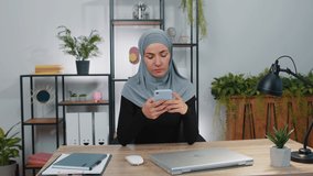 Young muslim business woman wearing traditional hijab headscarf in modern home office workspace texting share messages content on smartphone social media applications online, shopping, food ordering