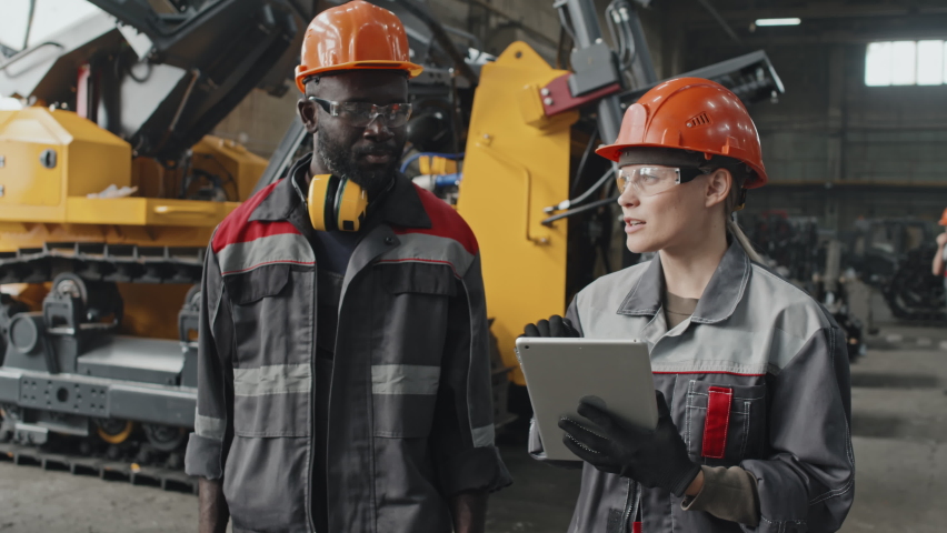 Interracial couple of technicians taking notes on tablet computer and having discussion while walking along tractor production plant Royalty-Free Stock Footage #1096656335