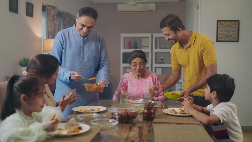 A happy Indian South Asian Ethnic Father son duo serving food at the dining table while rest of the Family including children, wife, mother, parents having lunch or a meal together in an indoor house. | Shutterstock HD Video #1096658755