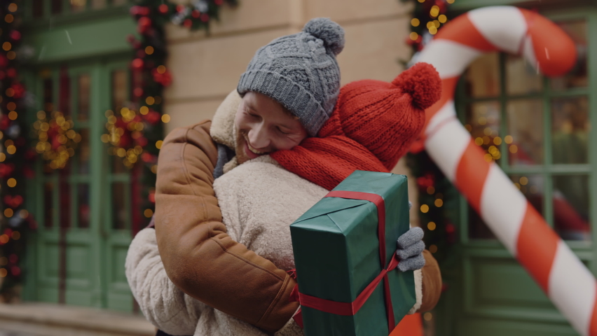 Happy smiled husband is going and giving his wife a christmas present standing on the decorated street. Hugs. Family tradition. Exchange of gifts