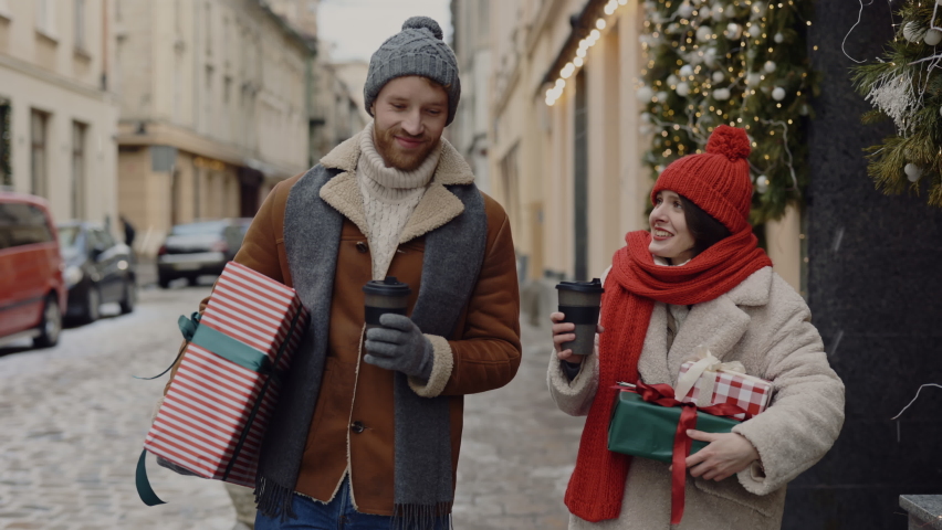 The caucasian couple is walking, lovely speaking and carrying christmas gifts together holding cups of coffee. The caucasian couple is going home from christmas shopping