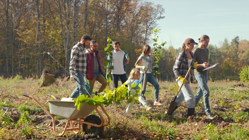 Happy females and males with kids ready for volunteering project with seedlings of trees in forest. Outdoors. Environmental volunteers in wood going to plant new trees on nice fall day. | Shutterstock HD Video #1096662587