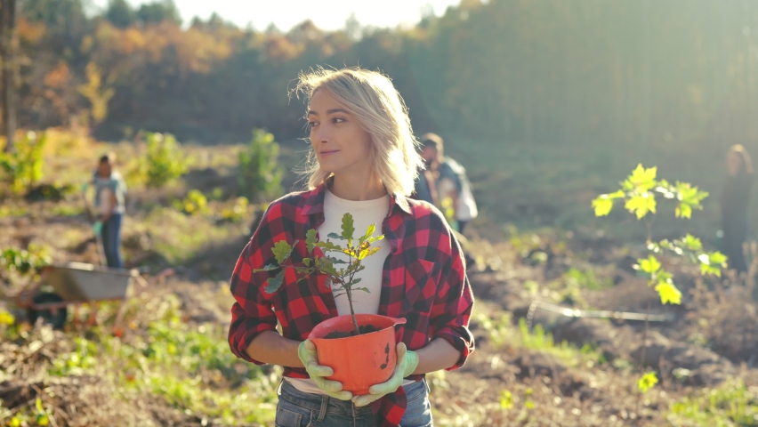 Portrait shot of blonde young beautiful Caucasian woman standing outdoor with tree seedling in pot and smiling cheerfully. Pretty female eco activist working against deforestation. Gardening. Royalty-Free Stock Footage #1096662697