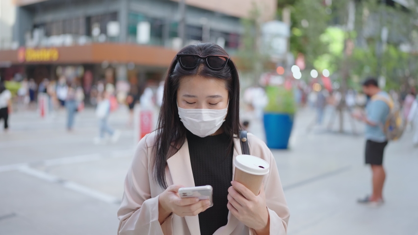 Asian woman wearing face mask, using smartphone and holding a cup of coffee on a city street