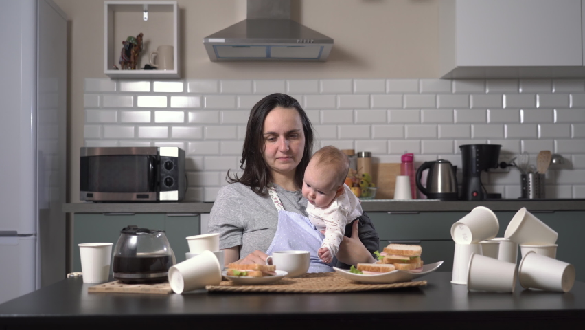 Exhausted young woman drinking coffee while holding her baby in hands in messy kitchen. Single mother and child caring concept | Shutterstock HD Video #1096669921