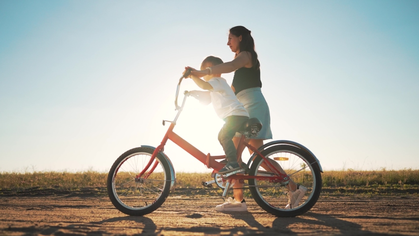 Happy family concept. Mom teaches her son to ride bike on green grass. Child rides bicycle along rural road. Green energy. Mom teaches her son to ride bike for the first time in park at sunset. Royalty-Free Stock Footage #1096672217