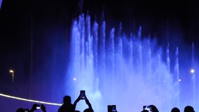 Crowd of spectators records a video on the phone of a beautiful bright performance of singing fountains at night. Grandiose musical show of fountains for the public is demonstrated late at night