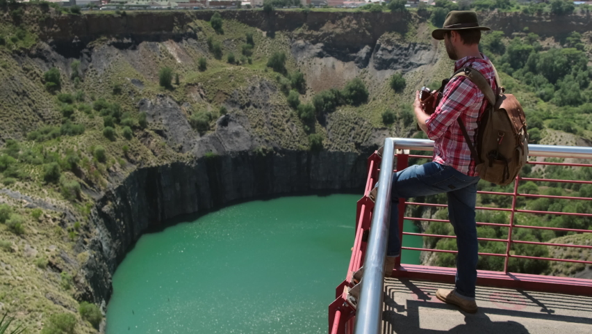 male traveler in a hat and a backpack makes a photo near a kimberlite pipe. Diamond quarry open pit top down view, on sunny summer day. Mining equipment for the transportation of minerals.