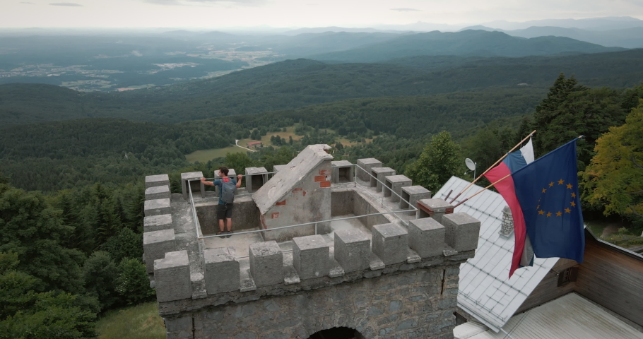 Drone shot of top of a tower where a young hiker with a backpack is taking a view in the valley. Slovenian and European flag fluttering in the wind. Cloudy day with a mid range view.