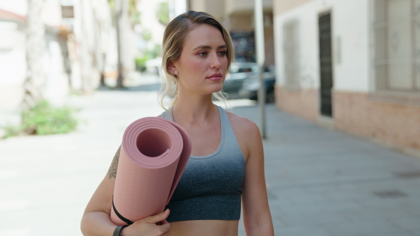 Young blonde woman wearing sportswear holding yoga mat at street