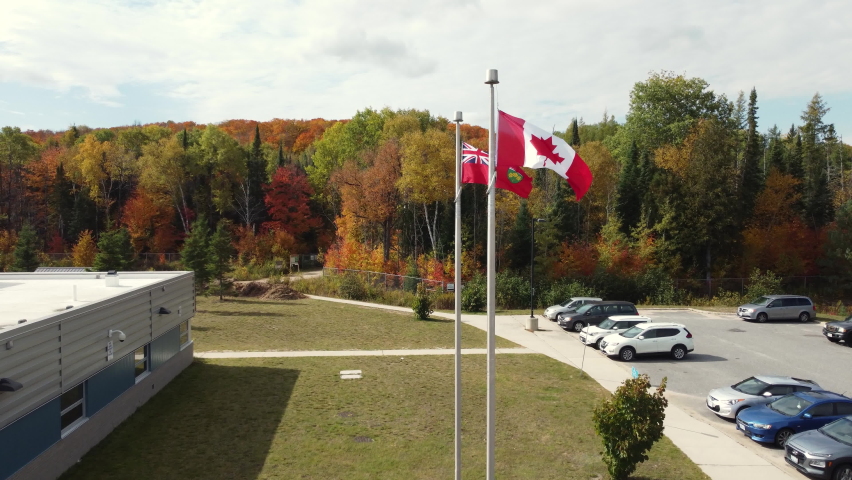 Flag Of Canada And Canadian Red Ensign Waving Outside Government Building In Muskoka Region, Ontario, Canada. Autumn Foliage In Background. pan left, drone shot