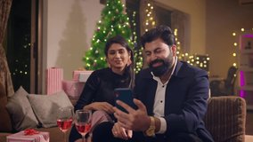 A happy smiling well dressed Indian Asian couple or husband and wife sitting together toasting wine glasses with family or friends talking on a video call using a mobile phone on Christmas festival