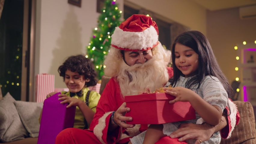 A happy smiling man or male dressed in a red Santa claus dress is talking and giving gifts to cute Indian Asian girl and boy or kids on the occasion of Christmas festival or holidays in house or home