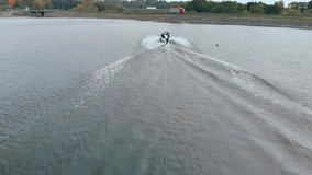 Drone Following Motor Boat Towing Wakeboarder on Scenic Lake in autumn. Aerial view (side) of woman rider wakeboard behind speedboat on desert body of water. Wakeboarder performs water jumps and trick