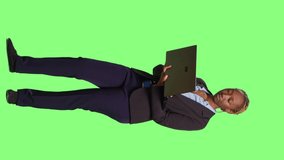 Vertical video: Company employee working on laptop over full body greenscreen background, wearing office suit and browsing internet website. Female worker using wireless computer to scroll through