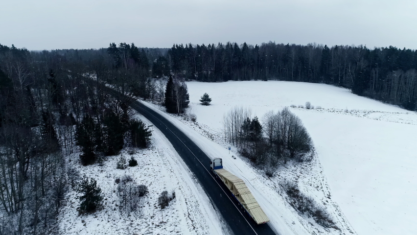 Aerial View Of Winter Snow Landscape With Semi Trailer Transporting Roof Trusses Along Road. Dolly Forward, Tilt Down