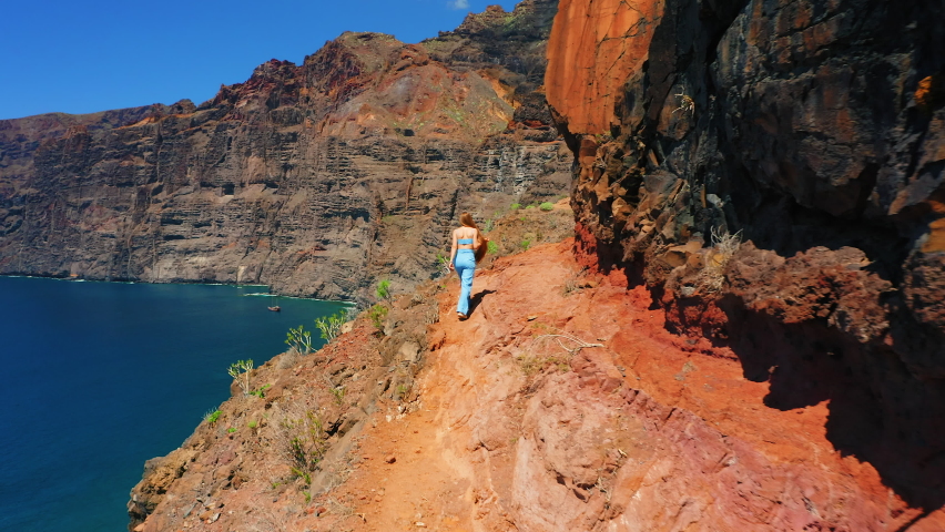 Young woman walkingon a steep slope. High sheer cliff sticking in open deep ocean. Beutiful girl tourist climbing a mountain observing the beauty of nature. Vacation on Los Gigantes Tenerife. Royalty-Free Stock Footage #1096728935