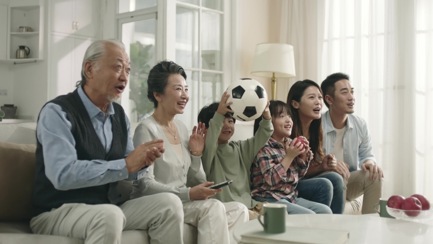 three generation asian family sitting on couch at home watching live broadcasting of soccer game together celebrating a goal Royalty-Free Stock Footage #1096730375