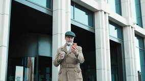Successful elegant grey-haired bearded man walking outside in city using looking at smartphone video calling holding cup of coffee. Stylish mature handsome serious senior male strolling on fresh air.