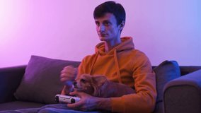 gamer playing video game on TV with modern console in neon light and holding his chihuahua dog