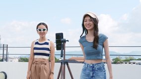 Young Asian woman with her friend er created her dancing video by smartphone camera together on rooftop outdoor at sunset To share video to social media application