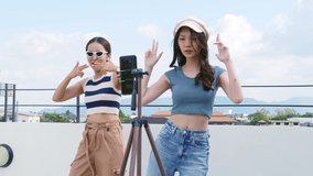 Young Asian woman with her friend er created her dancing video by smartphone camera together on rooftop outdoor at sunset To share video to social media application