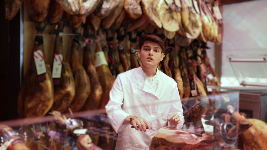  Confident positive young guy seller in white jacket standing in butcher shop, selling traditional spanish jamon Royalty-Free Stock Footage #1096743995