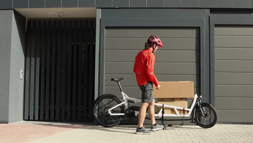 young courier with red clothing and helmet riding cargo bike arriving at the shipping destination to deliver a package to a city address, european housing, horizontal video	 Royalty-Free Stock Footage #1096750847