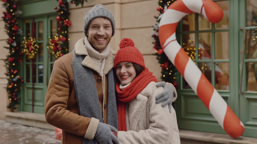 Smiled lovely caucasian couple standing in the street hugging and holding hands wearing winter warm clothes looking at the camera. Relationships and holidays concept