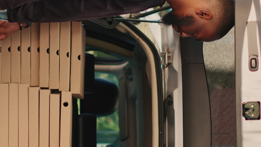 Vertical video: African american courier giving piles of pizza boxes to diverse people standing near building entrance. Food delivery employee delivering stacks of meal packages in car trunk. Tripod