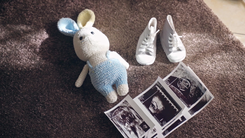 Ultrasound scan baby pictures, knitted bunny toy and baby shoes on the floor in sunlight, parents preparing for childbirth at home, enjoying period for pregnant woman. Maternity and pregnancy concept Royalty-Free Stock Footage #1096761575