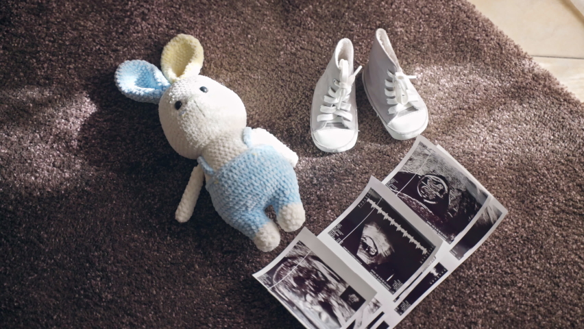 Ultrasound scan baby pictures, knitted bunny toy and baby shoes on the floor in sunlight, parents preparing for childbirth at home, enjoying period for pregnant woman. Maternity and pregnancy concept Royalty-Free Stock Footage #1096761575
