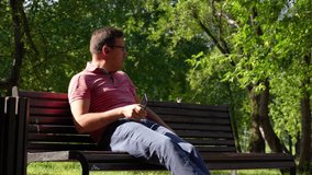 Carefree millennial man in eyeglasses is resting on bench while texting at mobile phone at city park