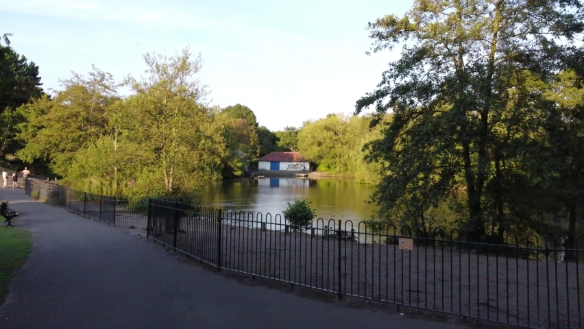 Low ariel drone view parkland lake wading birds. Landscape has wooded forestry area trees bushes stone building shed. Beautiful scenic sunny day sunshine on calm water blue sky above captured in 4k.