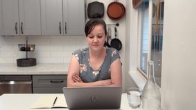 A woman working from home giving a presentation in her kitchen on her laptop wearing a flower patterned blouse