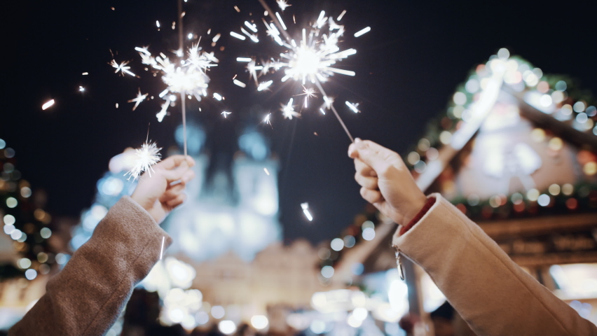 Celebrating New Year Christmas holidays vacation. Friends hands closeup with sparklers in air at decorated town square. Xmas Christmas Eve winter atmosphere. Unrecognisable people holding lights wave. | Shutterstock HD Video #1096782325